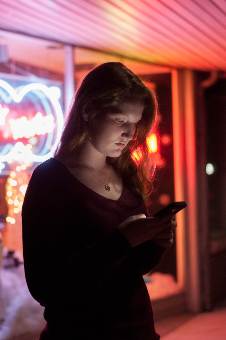 A woman stands near a neon sign. She stares at her phone.