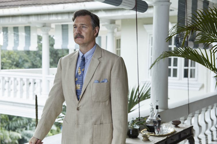 Mustachioed man in linen suit on varandah.