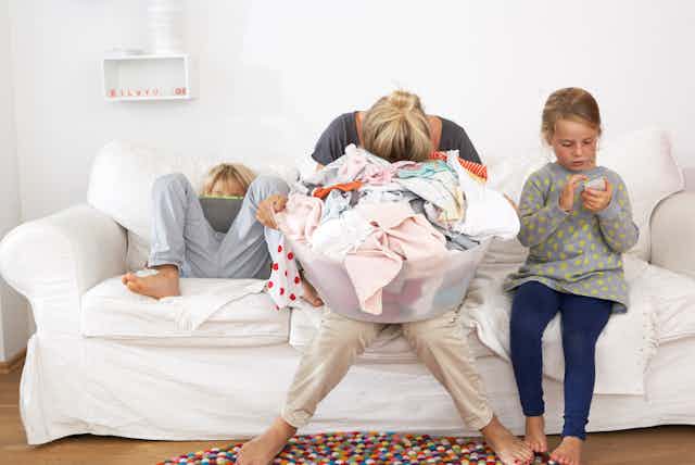 Exhausted mother with laundry basket on couch with children using digital tablet and cell phone.