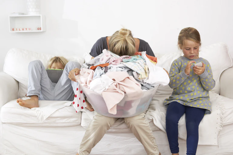 Exhausted mother with laundry basket on couch with children using digital tablet and cell phone.
