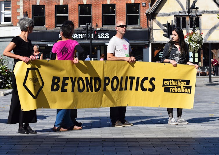 Four activists hold a banner reading 'Beyond Politics - Extinction Rebellion' in a town square.