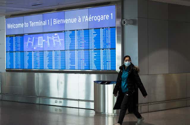 A woman wearing a mask walks past an arrivals sign at Pearson airport.