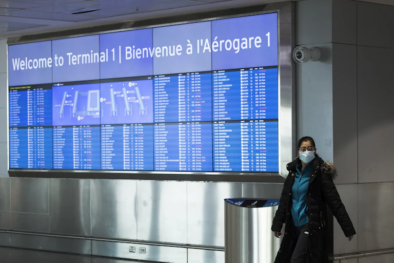 A woman wearing a mask walks past an arrivals sign at Pearson airport.