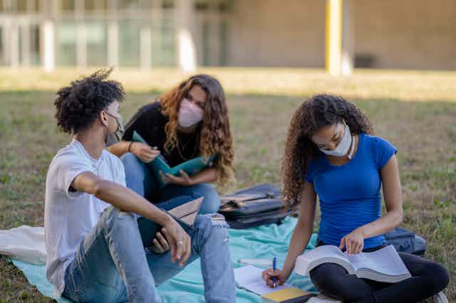 Three college students wearing masks study outside on a grass plain.