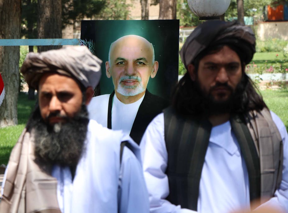 Two Taliban stand in front of a poster of Afghan president Ashraf Ghani