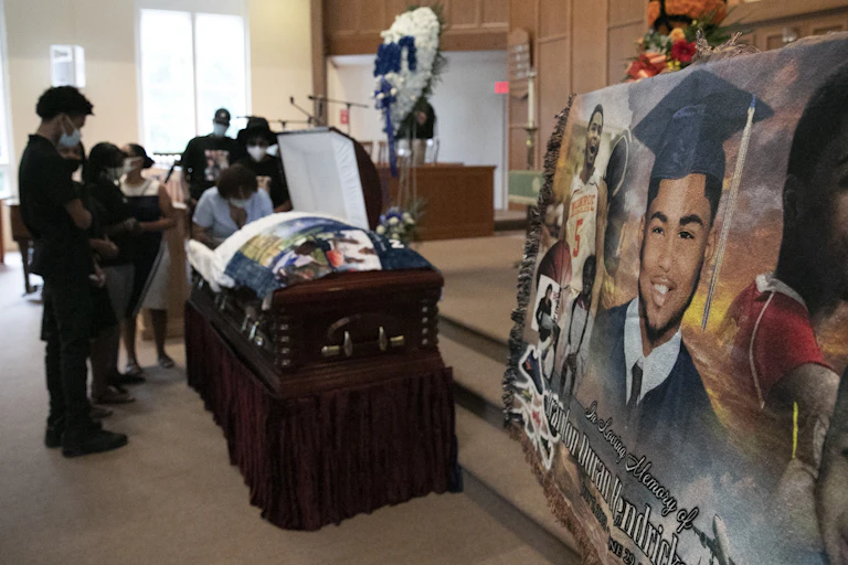 Mourners stand over the casket of 17-year-old Brandon Hendricks-Ellison while a photo montage of the 17-year-old is in the foreground.