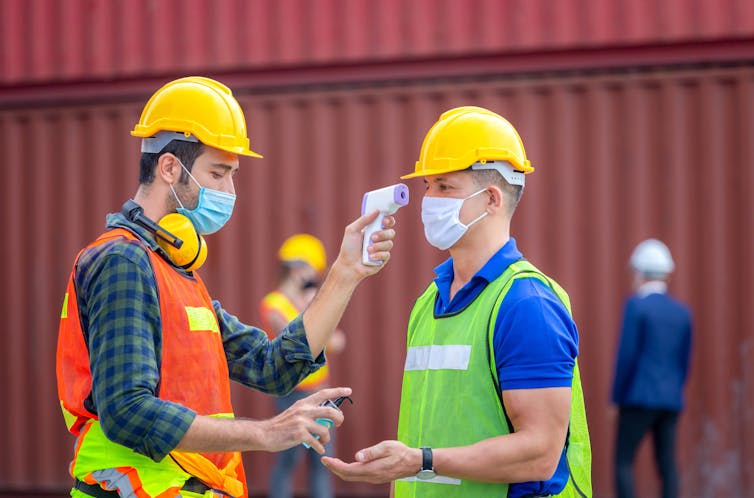 Factory worker takes temperature of colleague and holds out hand sanitiser.