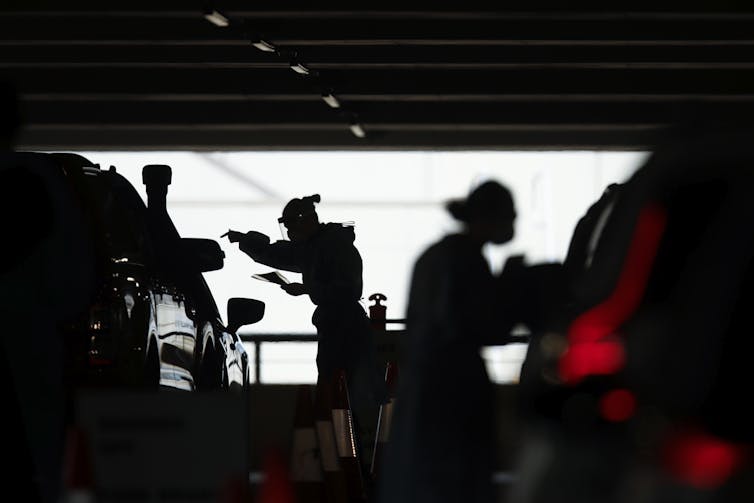 Two figures in PPE in silhouette lean towards cars.