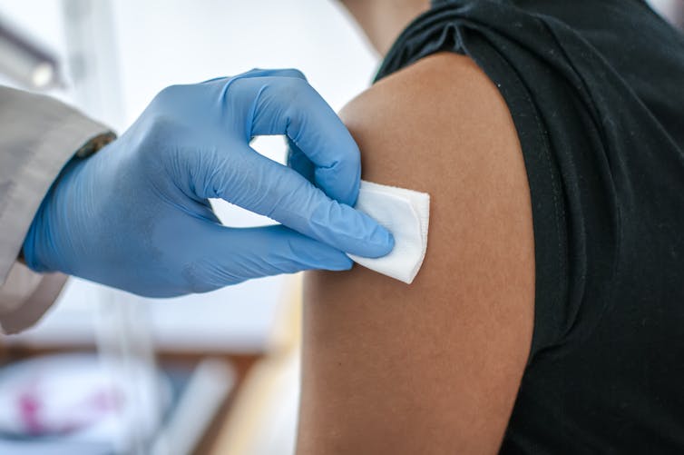 A doctor's gloved hand disinfects the skin of a patient's upper arm in preparation for a vaccination.