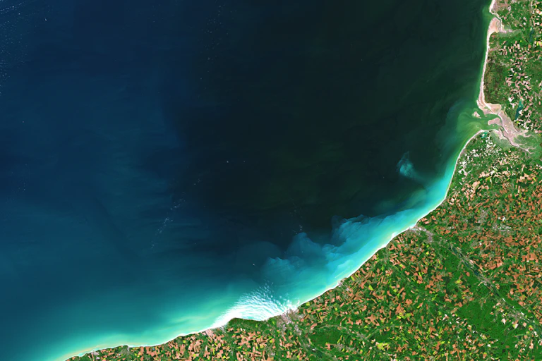 Vue du ciel, végétation et habitations sur la côte, mer et sédiments dans l'eau.
