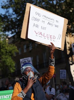 A woman holds a sign reading '540 colleagues to COVID-19 and we are not valued!' at a protest.
