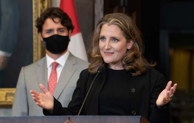 Chrystia Freeland gestures while talking to the media with Justin Trudeau standing behind her wearing a mask.