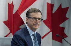 A man wearing glasses with Canadian flags as a backdrop.