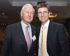 Two men in suits, one grey-haired and the other wearing glasses, stand smiling for the camera.