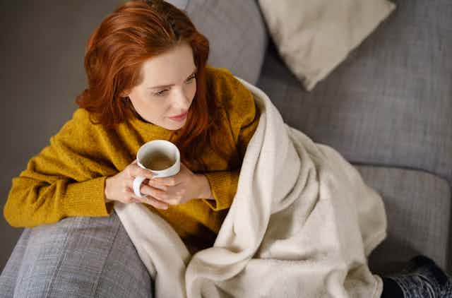 Young woman sitting on sofa wrapped in a blanket drinking a mug of tea