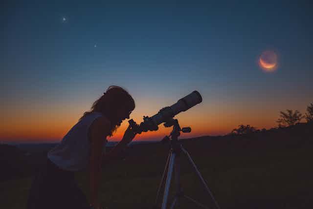 Silhouette of woman looking through a telescope at dusk.
