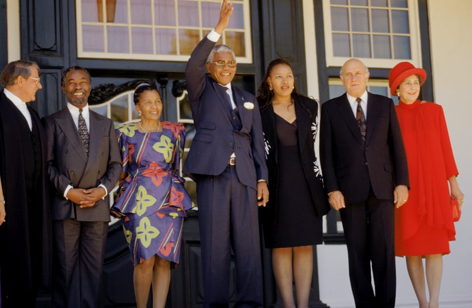 A grey-haired Nelson Mandela wearing a navy blue suit and specs waves his right hand, flanked by  Apartheid-era Justice Minister Kobie Coetsee, Thabo and Zanele Mbeki,  Zenani Mandela as well as  FW and Marike de Klerk.