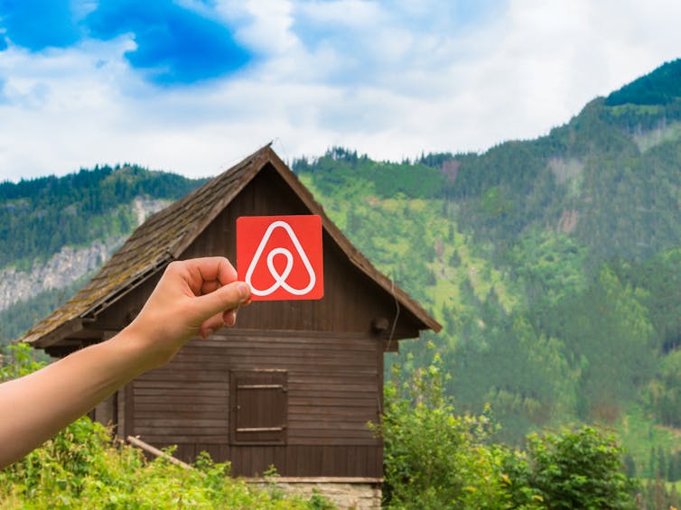Airbnb logo held by a hand in front of wooden hut in countryside.