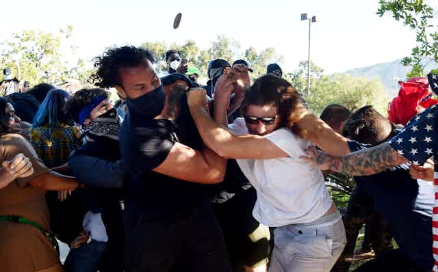Numerous people wearing masks in a physical clash with an unmasked man and another man wearing an American flag shirt 