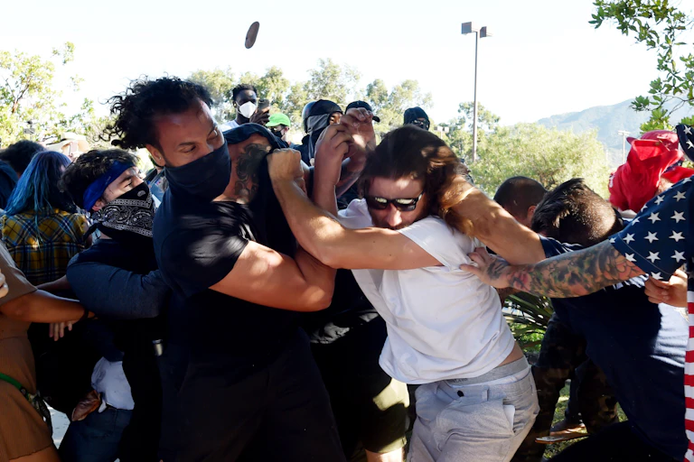 Numerous people wearing masks in a physical clash with an unmasked man and another man wearing an American flag shirt