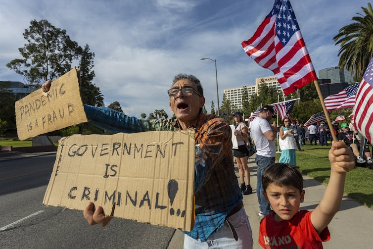 A man holds a sign saying 'government is criminal' and 'pandemic is a fraud' next to a child waving an American flag