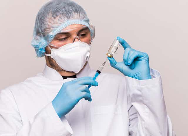 A man in a mask and lab coat filling a syringe with fluid