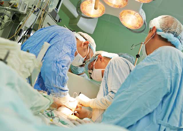 Three surgeons in blue scrubs work on a patient.