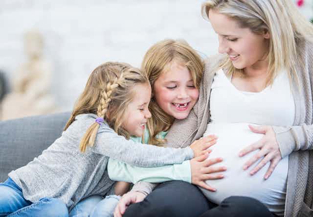 A pregnant white woman sitting on a sofa with two young girls who are smiling and touching her abdomen.