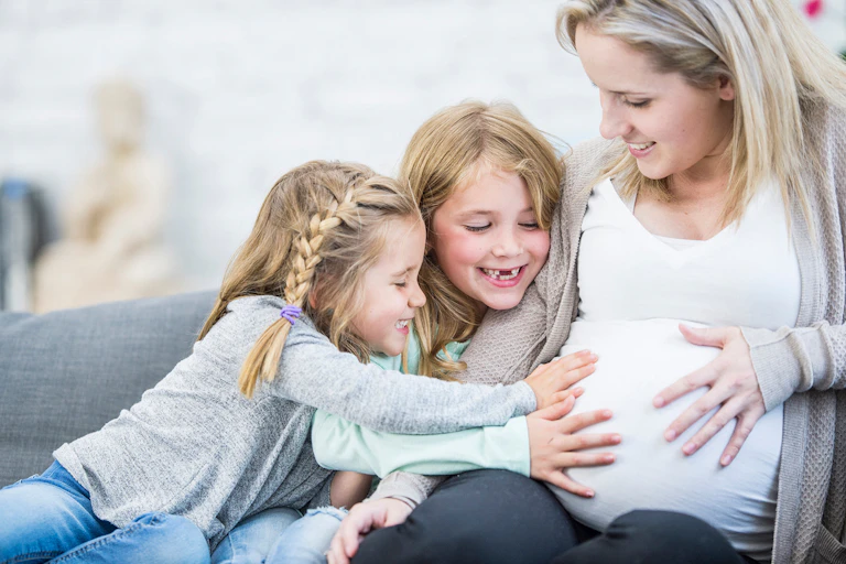 A pregnant white woman sitting on a sofa with two young girls who are smiling and touching her abdomen.