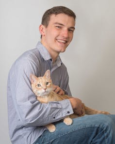 A smiling young man holds a cat in his lap.