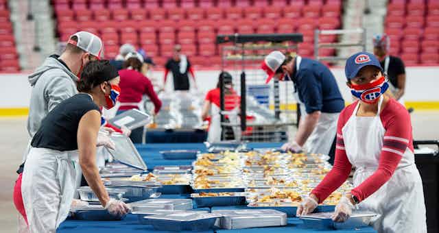 Four people wearing Montréal Canadiens face masks prepare containers of food with the red seats of a hockey arena behind them.