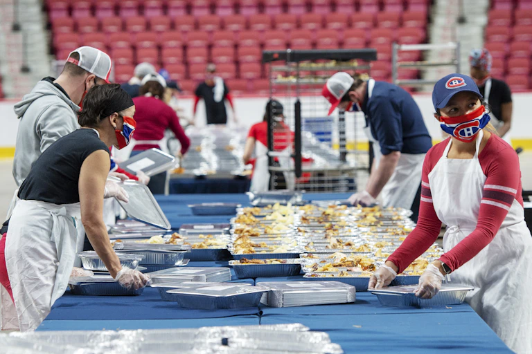 Four people wearing Montréal Canadiens face masks prepare containers of food with the red seats of a hockey arena behind them.