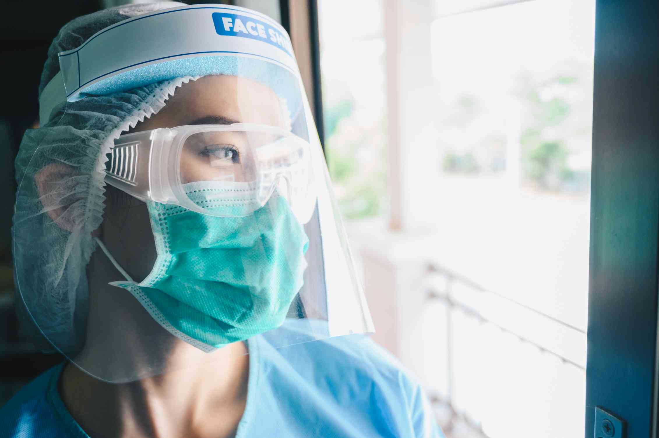 A health-care worker looking out the window. She wears full PPE, including a face shield.