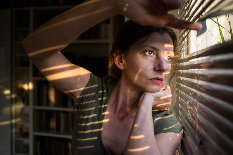 Young women in dark room looking out through venetian blinds