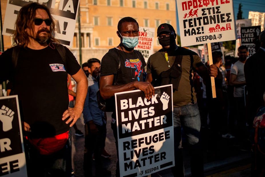 A protester wearing a mask holds a placard reading "Black Lives Matter, Refugee Lives Matter" during a demonstration. 