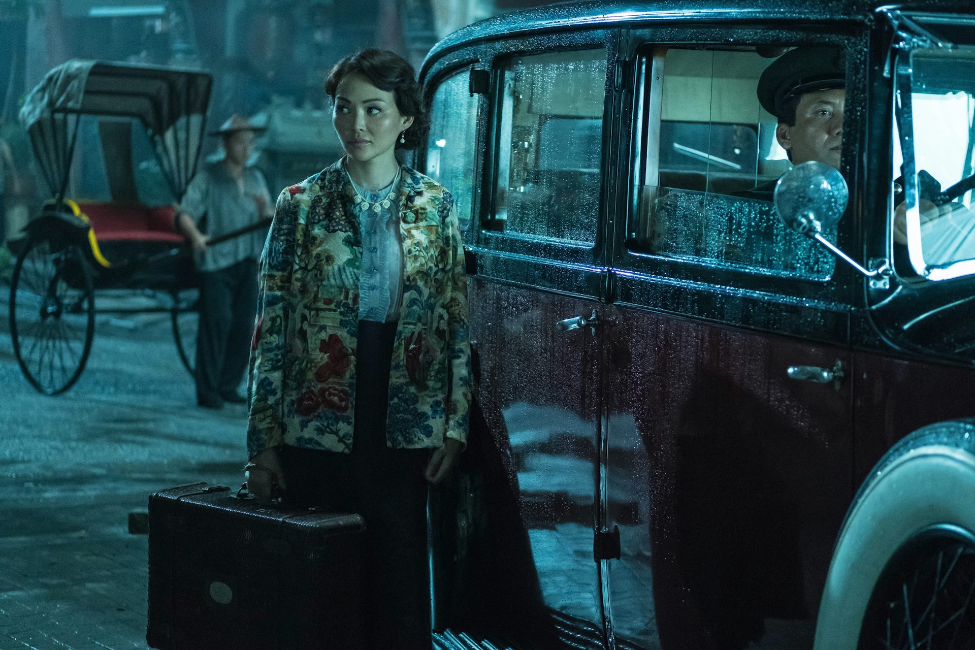 Woman stands with a suitcase next to an old fashioned car.