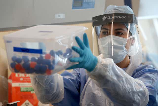 A nurse wearing PPE holds up a transparent box of coronavirus test samples. 
