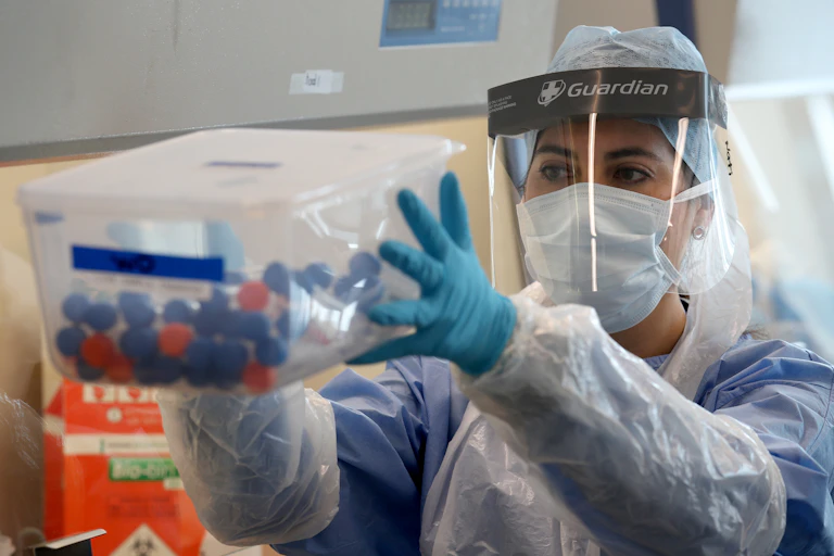 A nurse wearing PPE holds up a transparent box of coronavirus test samples.