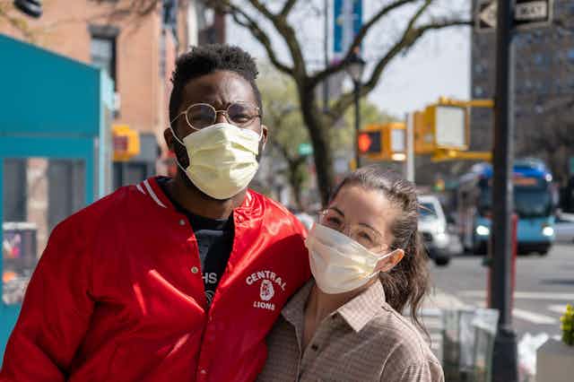 A man and woman wearing face masks pose for a picture on a sidewalk. 