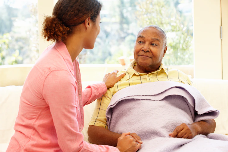 A young black woman holding the hand of an older black man with a blanket over him.