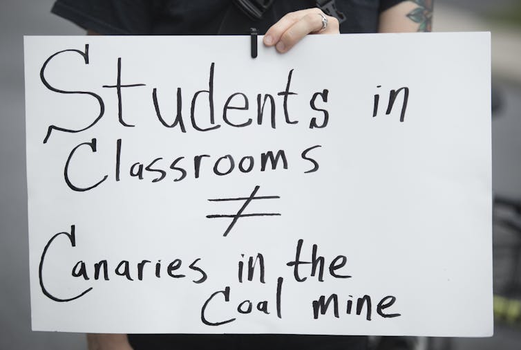 A protester holds up a sign that says Students in Classrooms do not equal Canaries in the Coal Mine