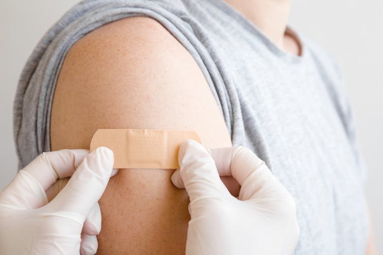 Gloved hands place a band-aid on a person's arm where they received a vaccination.