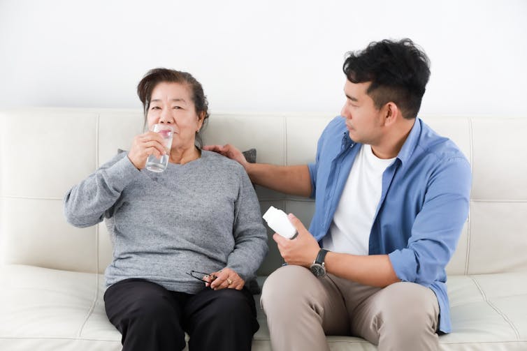 A young Asian man helping his mother with her medication. He holds a bottle of pills while his mother drinks from a glass of water as they sit side-by-side on a sofa.