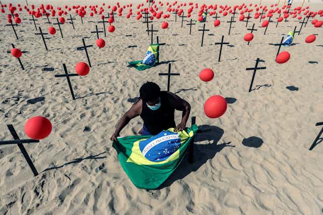 Man kneels with Brazil flag surrounded by an installation of balloons and crosses on the beach.