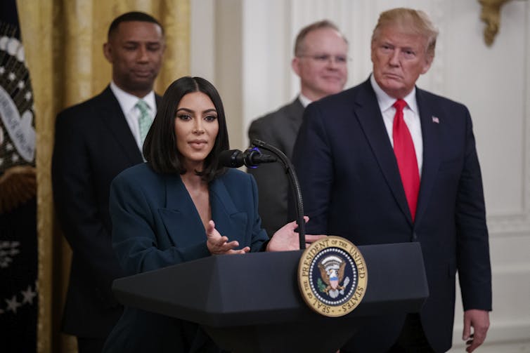 Kim Kardashian in a green suit speaking at a podium next to Donald Trump.