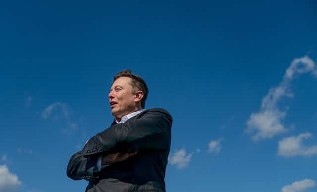 Elon Musk with arms folded, stood in front of blue sky.
