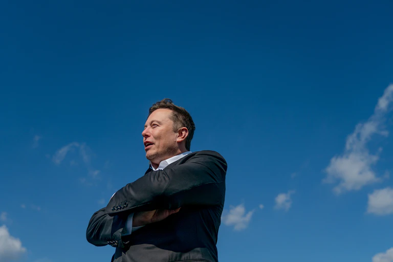 Elon Musk with arms folded, stood in front of blue sky.