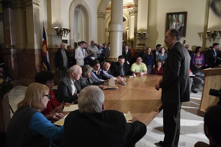 Electors sit around a large wood table in a stately setting, signing documents