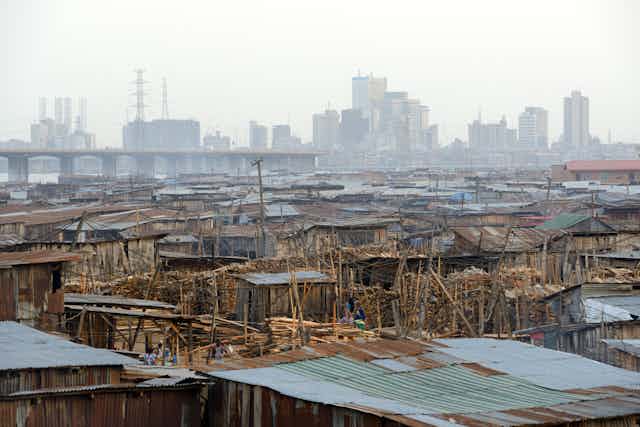 Shanty houses from wood and zinc in the foreground, sky-scraper buildings in the background, a bridge between them,
