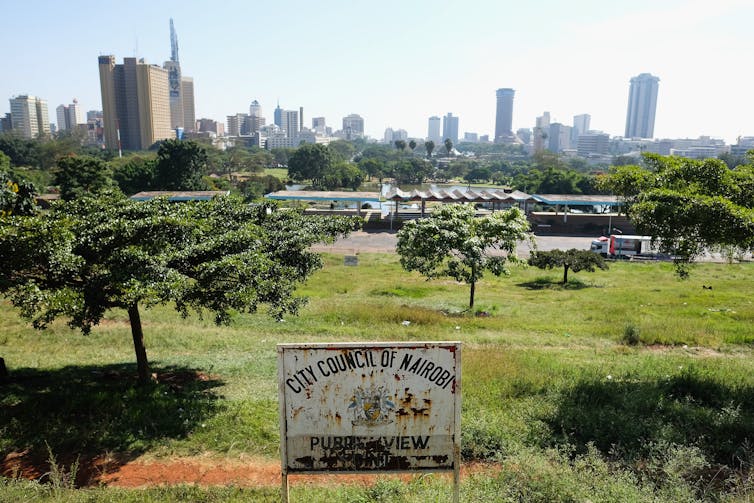 A rusty signboard for Nairobi city in the foreground with a vast green park with trees behind it and the cityscape in the distance with high-rise buildings.